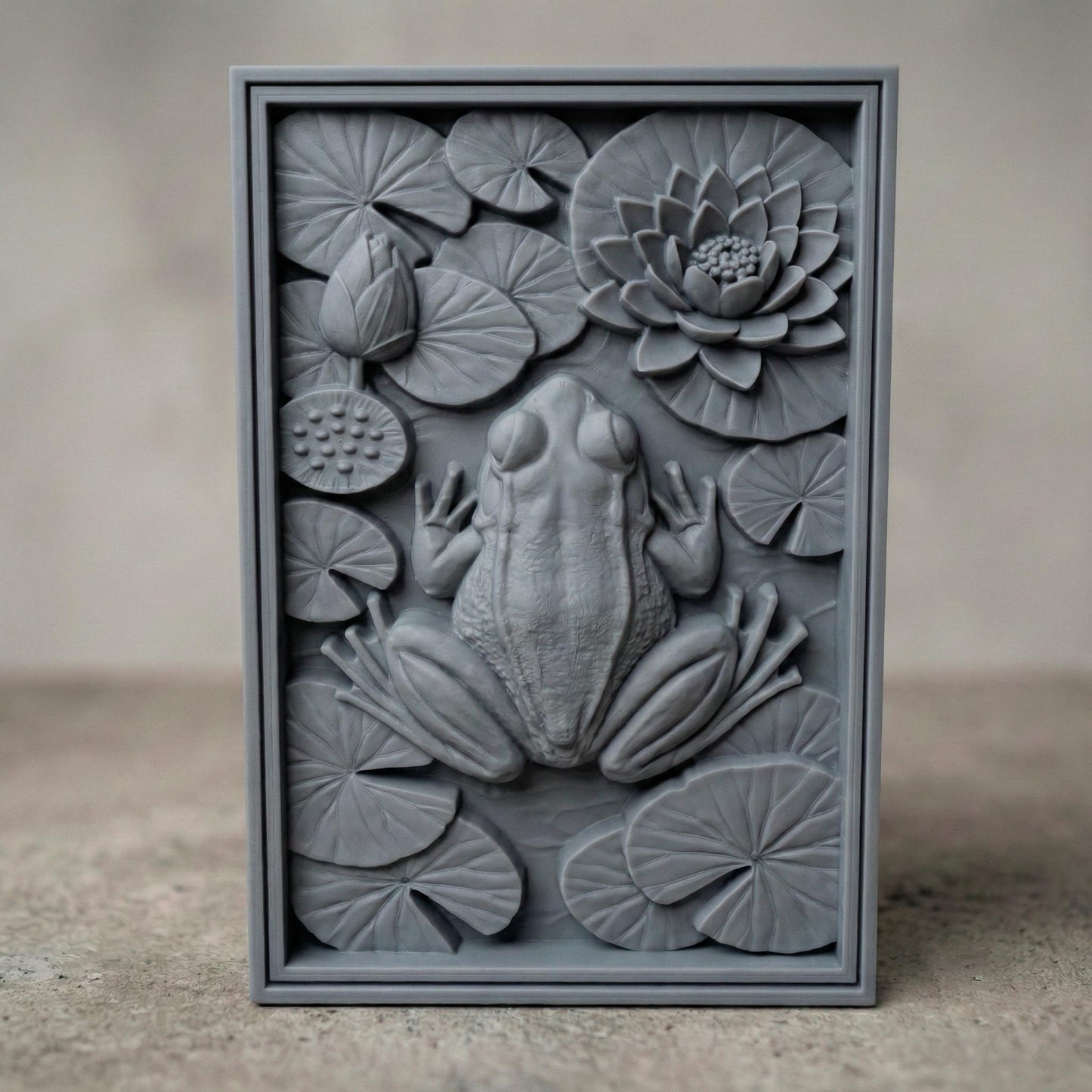 Detailed bas-relief panel showing a realistic frog resting among layered lily pads and a blooming lotus flower, symbolizing spring renewal and natural harmony. The composition features textured leaves, lotus seed pod, and overlapping circular pads creating rich depth and shadow. The frog’s sculpted back and limbs add lifelike dimension, ideal for CNC carving, 3D printing, and spring-inspired botanical wall décor.