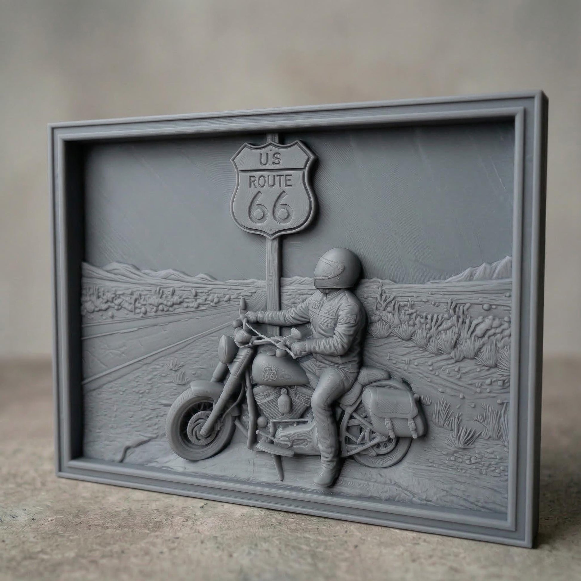 Detailed bas-relief scene of a motorcycle rider parked beside a US Route 66 road sign in a desert highway landscape. The rider wears a helmet and leather riding gear while sitting on a classic touring motorcycle with saddlebags. The background shows an open road stretching through desert terrain with cacti and distant mountains, symbolizing freedom, adventure, and the iconic American road trip culture.