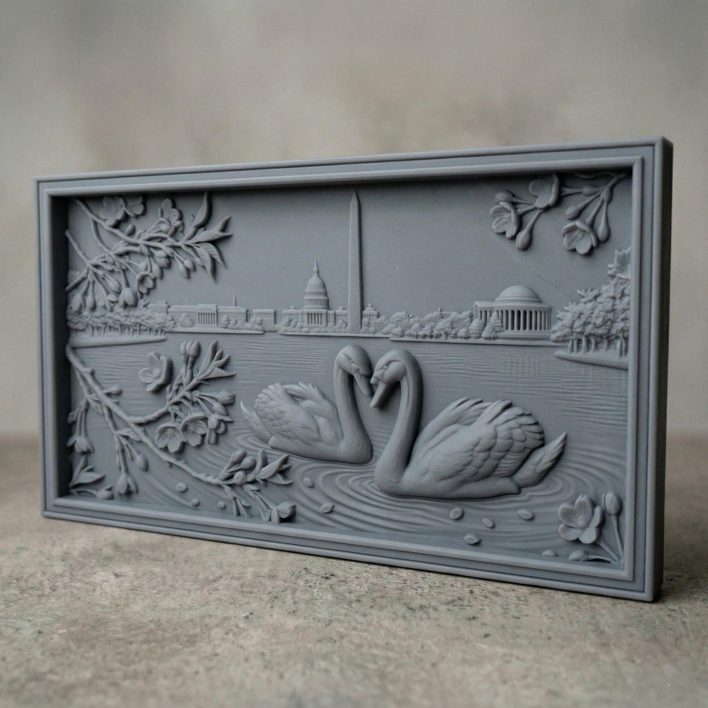 Detailed bas-relief carving of two swans forming a heart shape on rippling water, surrounded by blossoming branches. In the background, the Washington Monument and Capitol building rise along a serene lakeside skyline. Fine wood grain texture and layered depth create an elegant, romantic composition symbolizing love and unity in a peaceful natural setting.