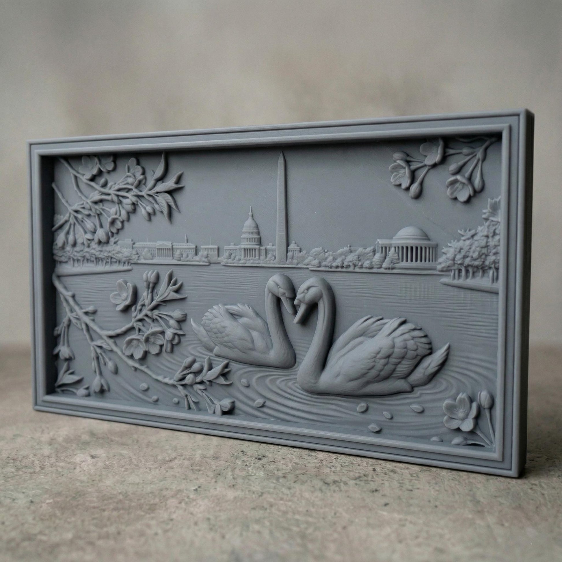 Detailed bas-relief carving of two swans forming a heart shape on rippling water, surrounded by blossoming branches. In the background, the Washington Monument and Capitol building rise along a serene lakeside skyline. Fine wood grain texture and layered depth create an elegant, romantic composition symbolizing love and unity in a peaceful natural setting.