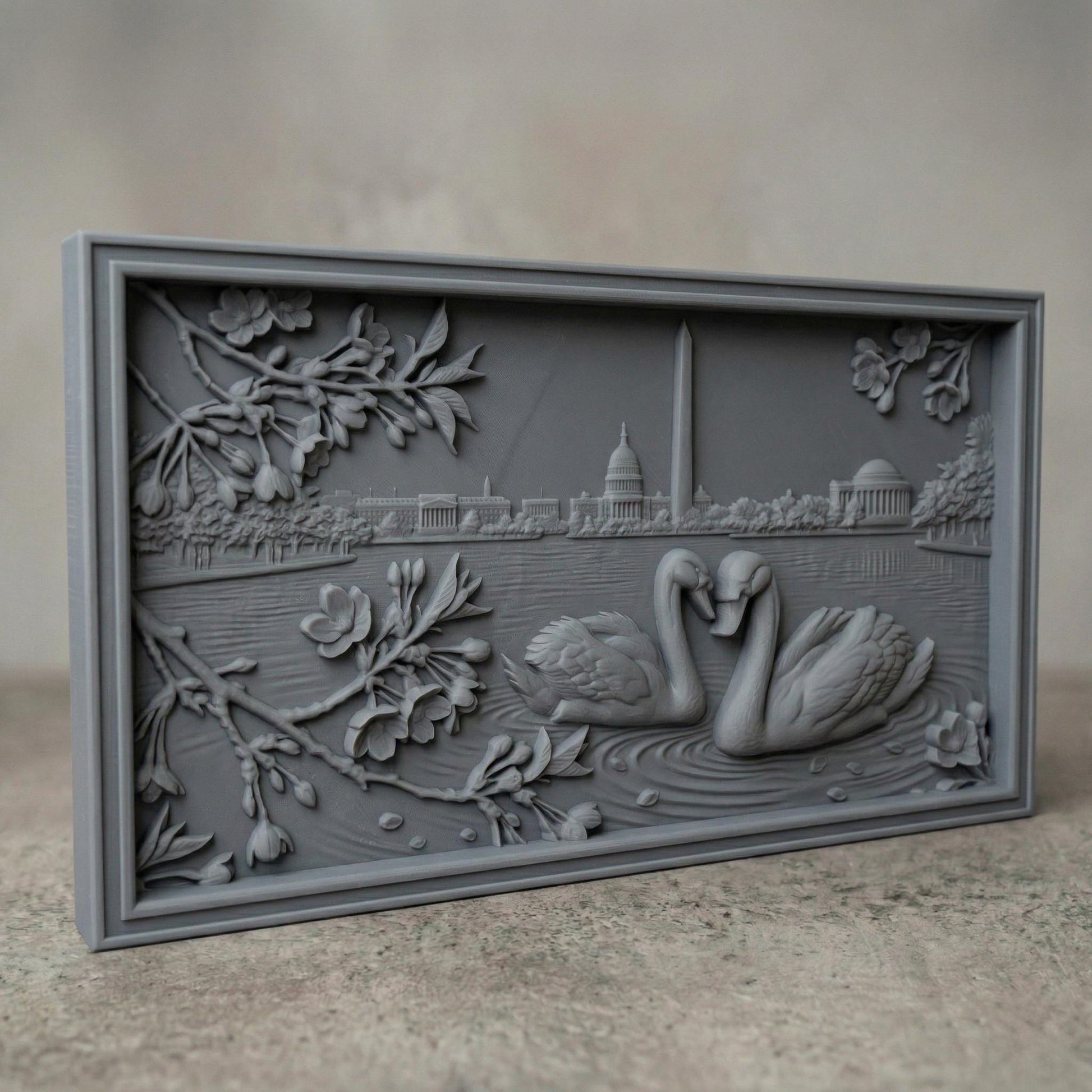 Detailed bas-relief carving of two swans forming a heart shape on rippling water, surrounded by blossoming branches. In the background, the Washington Monument and Capitol building rise along a serene lakeside skyline. Fine wood grain texture and layered depth create an elegant, romantic composition symbolizing love and unity in a peaceful natural setting.
