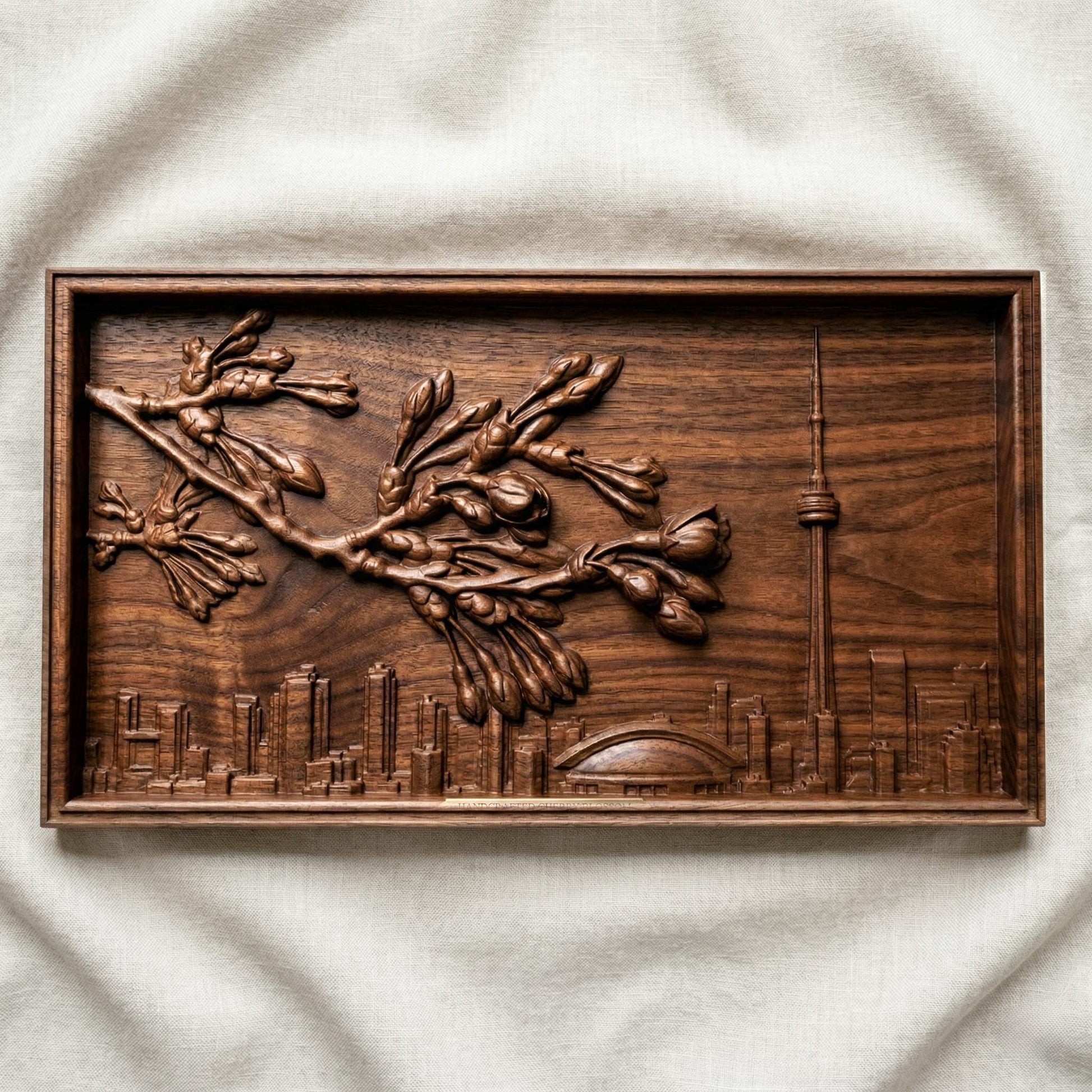 Elegant bas-relief carving of Toronto skyline featuring the CN Tower and Rogers Centre beneath a detailed cherry blossom branch in full spring bloom. The blossoms stretch across the scene above layered city buildings, with rich wood grain texture and smooth depth creating a harmonious blend of urban architecture and natural beauty.