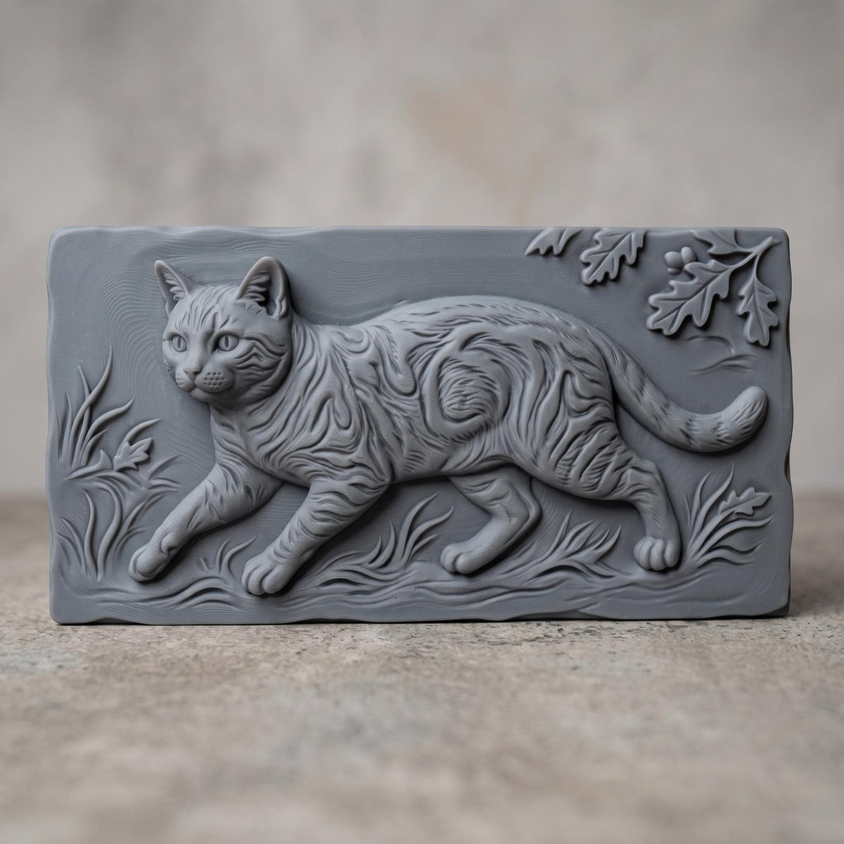 A tabby cat prowling through tall grass, carved in deep 3D bas-relief with fine fur striping, whisker detail, and expressive eyes. An oak leaf and acorn branch drapes across the upper corner, framed within a rectangular wood-textured panel with a naturally beveled edge. Designed for CNC relief carving, wood routing, and 3D printing as rustic wall decor.