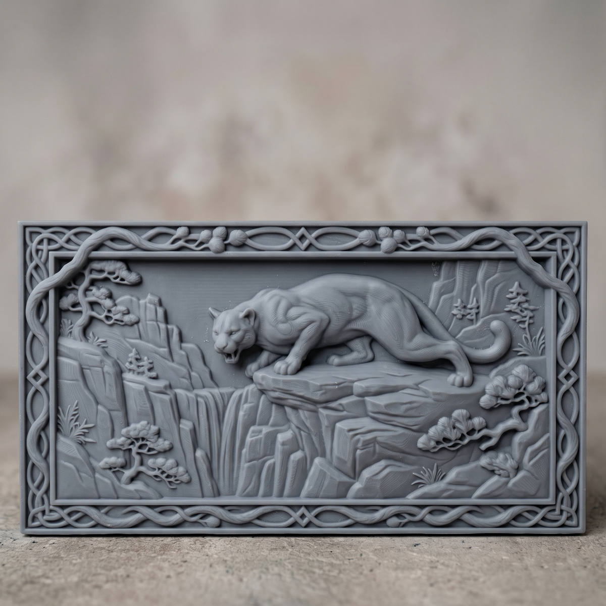 3D bas-relief of a mountain lion crouched on a rocky cliff edge above a waterfall, set in a landscape rectangular frame with a carved vine and Celtic knot border with pine cones. Bonsai-style pine trees, layered cliffs and grass surround the cougar. Smooth sculpted muscle detail and fine rock texture. Designed for CNC relief carving, woodworking and 3D printing as wide wall decor.